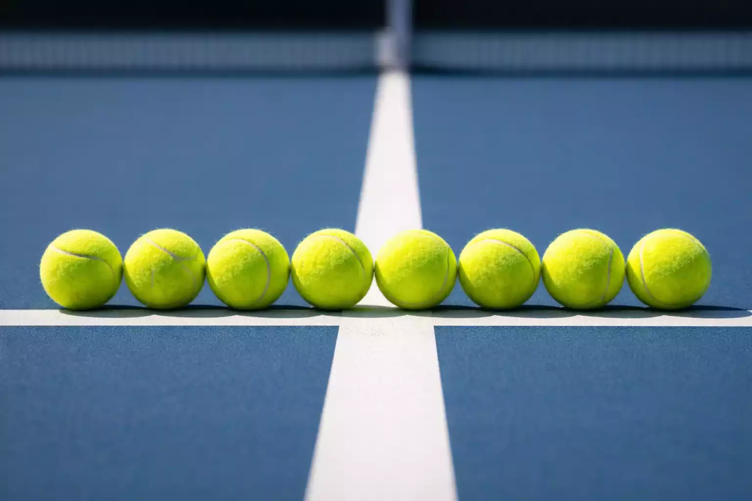 Varias pelotas de tenis alineadas sobre una pista dura azul junto a la línea blanca de la cancha en una composición ordenada